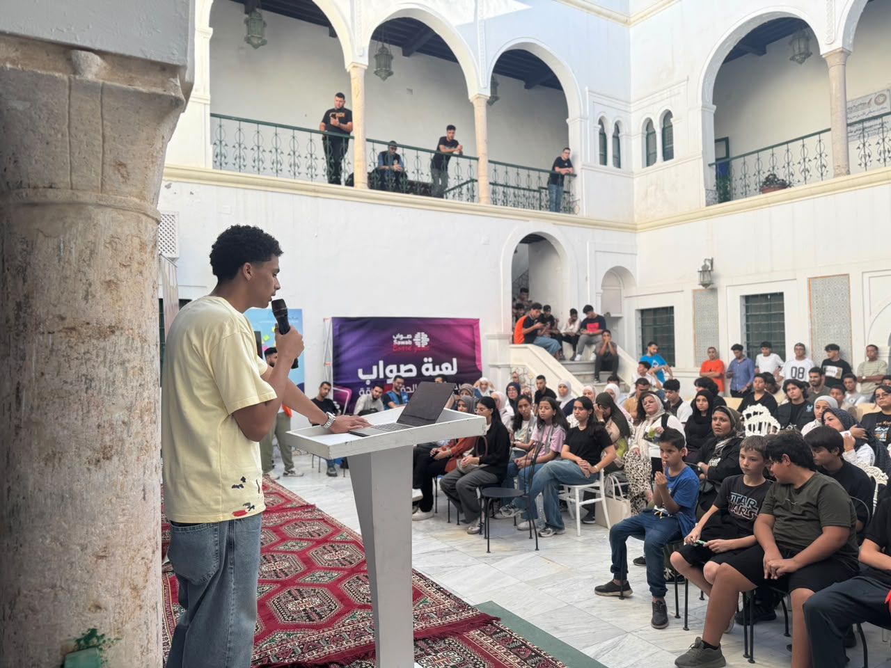 A student speaks into a microphone at a lectern in front of a full courtyard audience.
