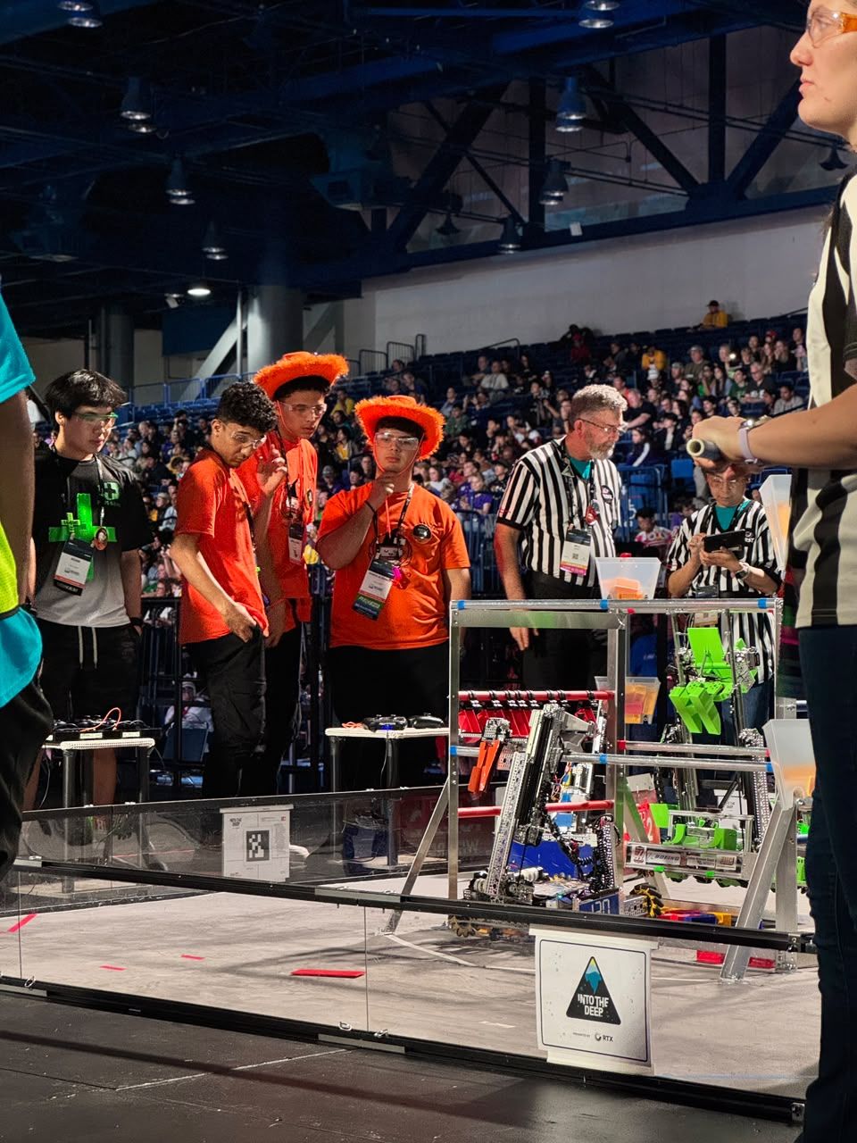 Drive team members in orange watch the field during an INTO THE DEEP match, with referees beside them.