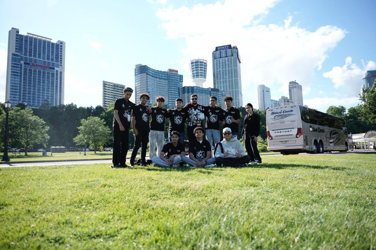 Team members in black team shirts pose together on a lawn, with tall buildings behind them.